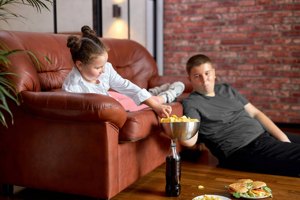 Fat Teenager Boy Sharing Crisps With Sister Sitting On Floor, Having Rest Together - Foto, immagini
