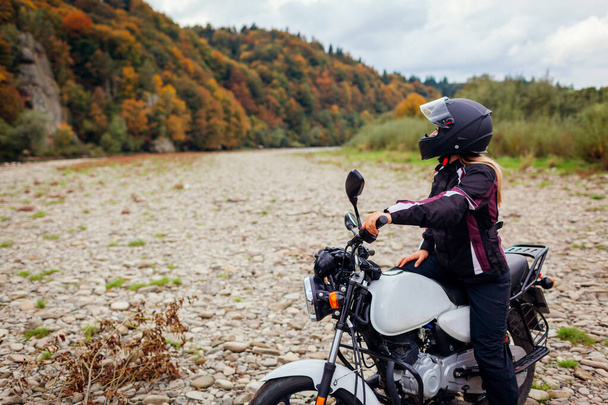 Woman biker travel by motorbike in fall. Motorcyclist enjoys autumn landscape in mountains having rest by forest. Traveler sitting on motorcycle wearing helmet, jacket - Photo, image