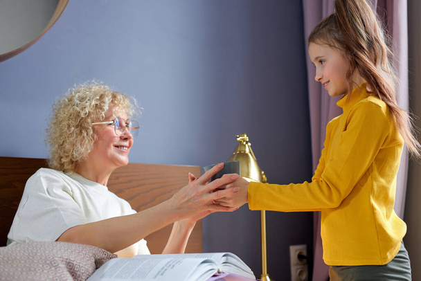 Caring child girl giving cup of tea to granny sitting on bed in cozy room, family love - Фото, зображення