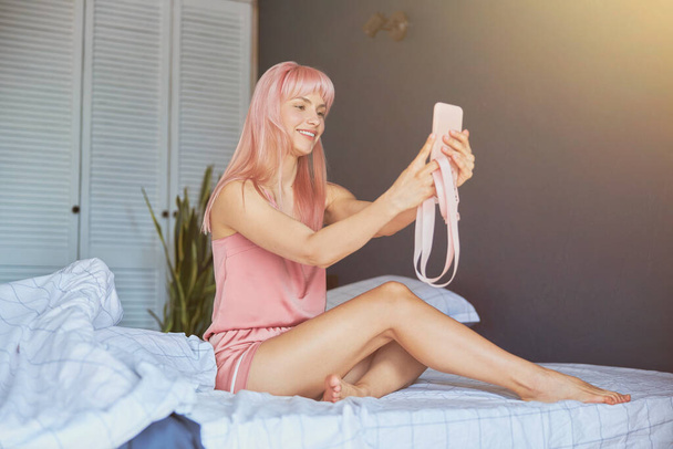 Спокуслива жінка з рожевим волоссям в елегантній шовковій піжамі бере селфі на велике ліжко
 - Фото, зображення