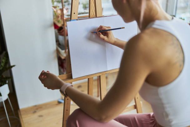 Woman artist draws picture on white paper with graphite pencil at wooden easel in light studio - Photo, Image