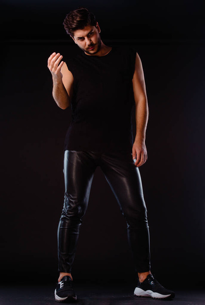 Cool young man dressed in black sleeveless t- shirt posing in studio agaisnt black backgorund - Photo, Image