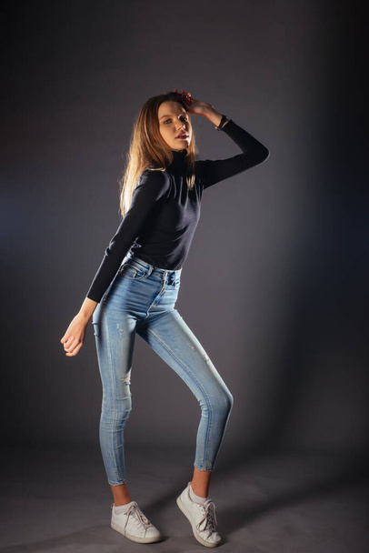 Full-length picture of a beautiful female model posing on dark background in a studio wearing a black blouse, jeans and white sneakers - Fotoğraf, Görsel