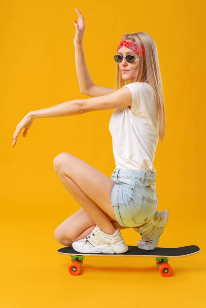 Skateborder κορίτσι με σορτς και T-shirt κάθεται στο πλοίο πάνω από κίτρινο φόντο - Φωτογραφία, εικόνα