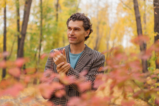 Tall handsome man dressed in a brown jacket walking in the autumn alley and posing near the bush with red leaves. - Fotó, kép