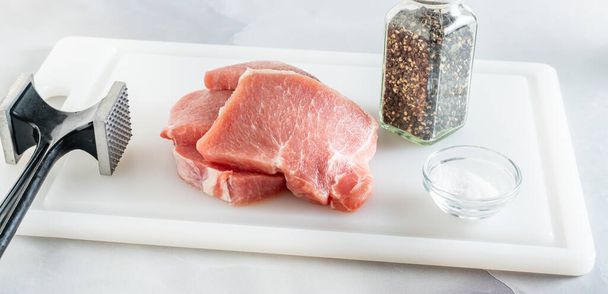 Rohe Schweinekoteletts und Fleischhammer mit Salz und schwarzem Pfeffer hautnah auf Schneidebrett - Foto, Bild