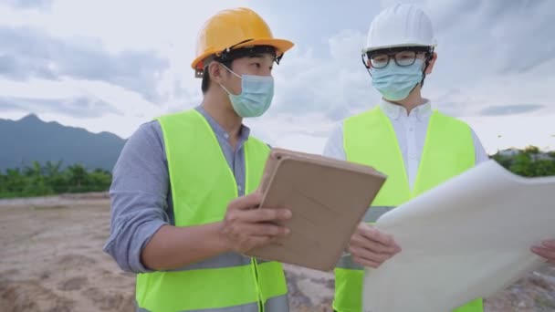 Los trabajadores de la construcción asiáticos usan mascarilla médica y uniforme de seguridad completo trabajando juntos durante la pandemia en el sitio de construcción al aire libre, sosteniendo la tableta con informe al gerente, proyecto ecológico  - Imágenes, Vídeo