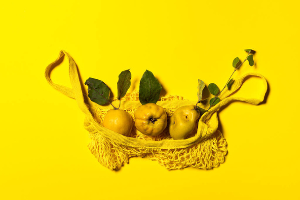 Grupa jabłek pigwy z naturalnymi liśćmi i gałązką w żółtej netto torbie zakupowej. Mieszkanie leżało na żółtym papierze. Owoce i liście z naturalnymi plamami i zadrapaniami. Orientacja pozioma. - Zdjęcie, obraz