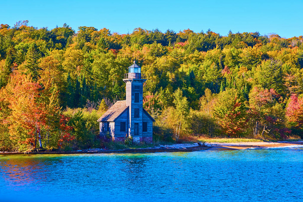 Old wooden lighthouse on edge of island with forest behind it and calm lake ocean or sea water in front of it - Photo, Image