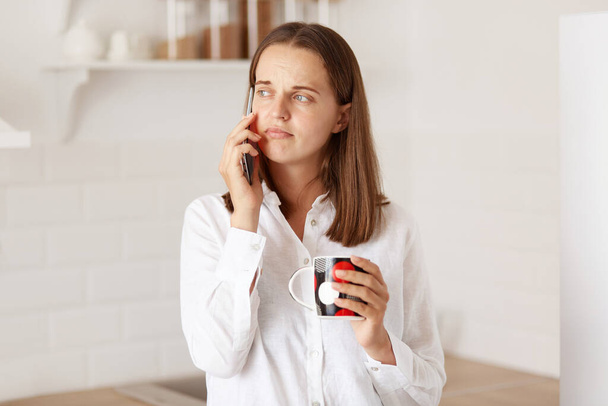 Portrait of sad pensive woman talking on the mobile phone, holding a cup, looking away with upset expression, posing in kitchen, wearing casual style closing. - Foto, Bild