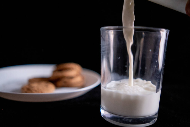 Versare il latte nel bicchiere accanto al piatto di biscotti con sfondo nero - Foto, immagini