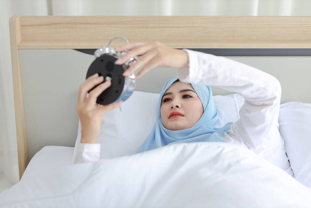 Nukkuva aasialainen musliminainen valkoisissa yöpuvuissa sängyllä, herätyskellorengas kadoksissa. Muotokuva nuori tyttö yrittää lopettaa soi aamulla - Valokuva, kuva