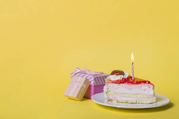 illuminated candle slice cake with two gift boxes against yellow background. High quality beautiful photo concept - Valokuva, kuva
