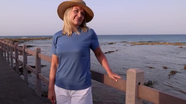 Smiling middle aged woman wearing blue t-shirt and straw hat and standing at sunset on the wooden bridge with hands on it. T-shirt mockup - Footage, Video