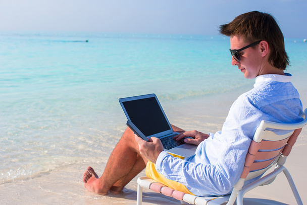 Joven hombre de negocios usando portátil y teléfono en la playa tropical
 - Foto, imagen
