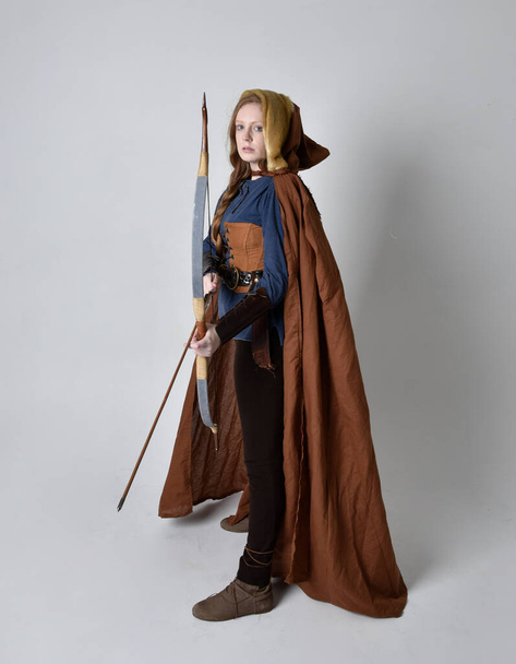 Longitud completa, retrato de mujer de pelo rojo con traje inspirado vikingo medieval y capa que fluye, sosteniendo arco un arma de flecha, posando sobre el fondo del estudio. - Foto, imagen