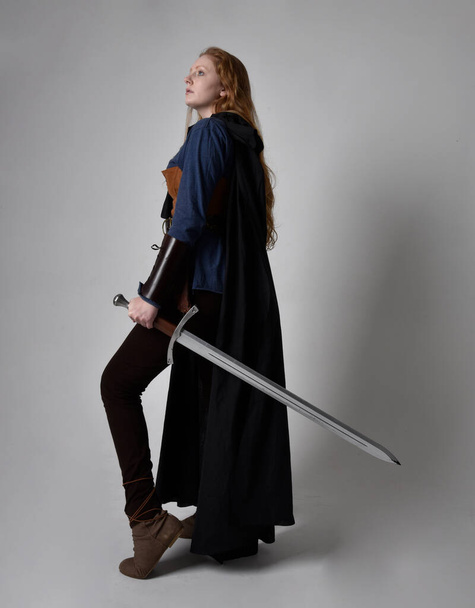 Teljes hossz, portré vörös hajú nő visel középkori viking ihletett jelmez és áramló köpeny, kezében egy hosszú kard fegyver pózol ellen stúdió háttér. - Fotó, kép