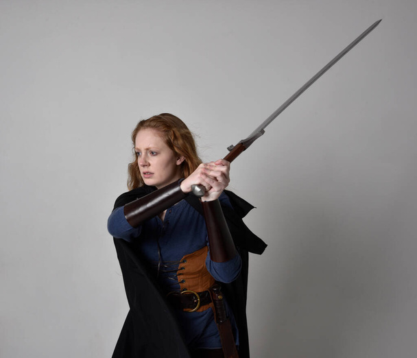 Retrato de cerca de la mujer de pelo rojo con traje inspirado vikingo medieval y capa que fluye, sosteniendo un arma de espada larga, pose de acción contra el fondo del estudio. - Foto, imagen