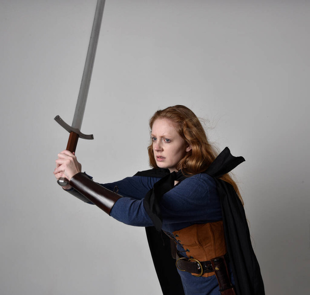 Крупный план портрет рыжеволосой женщины в средневековом костюме викинга и плаще, держа длинный меч оружие, действие поза на фоне студии. - Фото, изображение