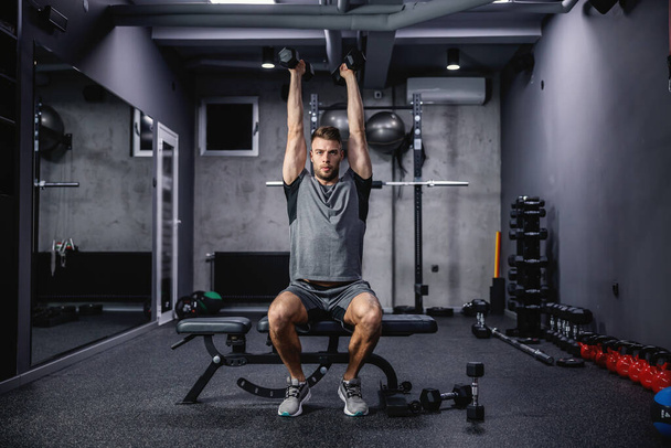 Slim and fit. Static weight loss training for a slim and fitness body. A sportsman focused on training sits on a bench in the gym and raises his dumbbells above his head. Fitness lifestyle, sport - Photo, Image