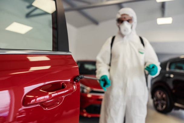 Mann in Schutzuniform mit Maske und Schutzbrille desinfiziert rote Autotür in einem modernen Autosalon-Showroom. Er reinigt Bereiche, die häufig berührt werden, verhindert eine Infektion mit dem Covid-19-Virus Coronavirus - Foto, Bild