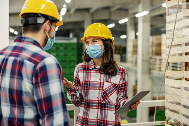 The female team leader smiles at her college as she prepares goods for transportation and delivery. On the digital tablet, she shows him the work plan in the factory warehouse. Covid19 stay healthy - Photo, Image