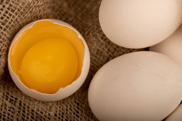 Een gebroken wit kippenei en een aantal hele eieren op een huiselijke stof met een ruwe textuur, close-up selectieve focus - Foto, afbeelding