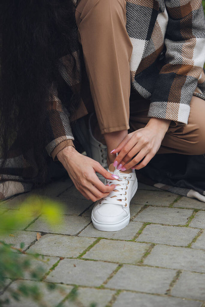 Closeup photo of young woman wearing checkered long coat, and beige pants . Lady posing on city street. Women ties laces on sneakers - Foto, Bild