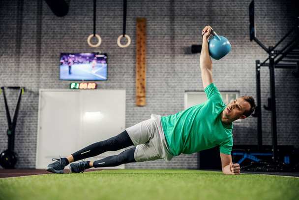 Kestävyys ja vahvistaminen koko kehon ja ydin lihaksia. Urheilullinen lihaksikas mies tekee sivulankun, jossa on sininen vedenkeitin toisessa kädessä, joka on nostettu korkealla päänsä yläpuolella. Urheilupukuinen mies pumppaa lihaksiaan - Valokuva, kuva