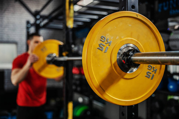 Preparando el peso de la barra para el entrenamiento fuerte de la quema del músculo. Un hombre en ropa deportiva roja monta pesas de pesas en el gimnasio. Primer plano de una barra amarilla en una barra. Fondo borroso - Foto, imagen