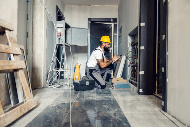 An experienced worker is kneeling in the hall of the building in the construction process and taking notes. In front of him are pipes for gas so, it has to be properly done. - Foto, afbeelding