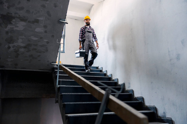 Un homme à tout faire barbu avec un casque sur la tête avec une boîte à outils dans les mains descend les escaliers dans un nouveau bâtiment dans le processus de construction. Il y a beaucoup de travail à faire. - Photo, image