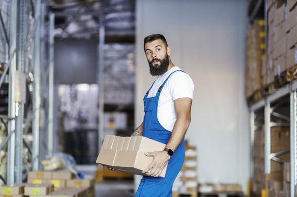 A strong, muscular warehouse worker in overalls is relocating boxes from one place to another. There are a lot of goods for shipping, so he must hurry up. - Foto, Imagem