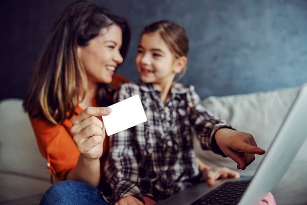Mother and daughter sitting on the sofa and using laptop for online shopping. Mother is holding credit card while daughter pointing at toy she wants. Selective focus on credit card. - Foto, Imagen