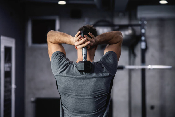 Shoulder and arm exercises. Rear shot of a handsome man holding a dumbbell between his shoulder blades and strengthening his arm, shoulder, and back muscles in modern sport center - Photo, Image