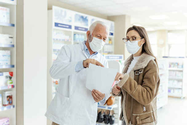 Reading prescription drugs, a conversation between pharmacist and client. A mature man explains drug therapy to a female client. They both wear protective masks due to the corona virus situation - Photo, Image
