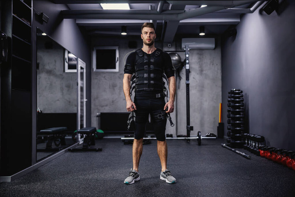 Ένας άνδρας με αυτοπεποίθηση εκπαιδευτής ντυμένος με ειδική στολή για την τεχνολογία EMS στέκεται στο γυμναστήριο με μια σκοτεινή γκρίζα ατμόσφαιρα. Επανάσταση στην κατάρτιση, την αποκατάσταση του σώματος, τους εργαζομένους και το προσωπικό γυμναστικής - Φωτογραφία, εικόνα