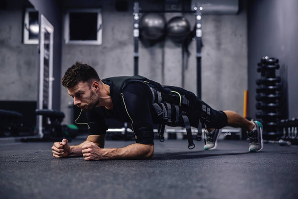 Yeni EMS teknolojisi kullanarak tüm vücudu yakma egzersizi. EMS için giysi giyen çekici genç bir adam spor salonunda dirseklerinin üzerinde tahta bir pozisyonda. Elektriksel kas uyarılması - Fotoğraf, Görsel