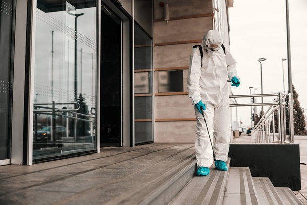 Ένας άνδρας με λευκή προστατευτική στολή και μάσκα απολυμαίνει την μπροστινή πόρτα και τις σκάλες του κτιρίου γραφείων με χημικά. COVID-19 πρόληψη της απολύμανσης, κορώνα, πανδημία - Φωτογραφία, εικόνα