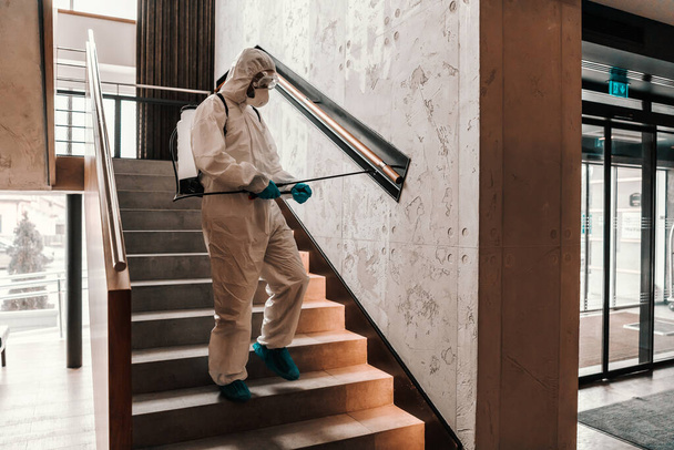 Προστασία του Covid19. Ένας άντρας με προστατευτική στολή αποστειρώνει τη σκάλα στην μπροστινή πόρτα του ξενοδοχείου. Προστασία του περιβάλλοντος, coronavirus, ασφάλεια ταξιδιού - Φωτογραφία, εικόνα