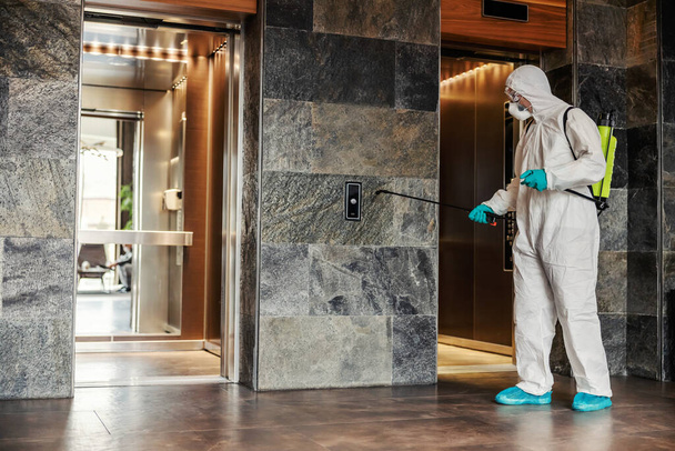 By disinfecting the space and air against the symptoms of coronavirus - fever, cough, headache. A man in a protective uniform cleans high-risk areas of infection such as elevator buttons - 写真・画像