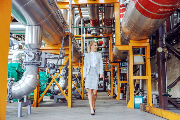 Smiling blond female power plant owner walking around and checking on machinery. Full length of hardworking blond female supervisor. A positive attitude of an adult woman in an elegant suit - Photo, Image