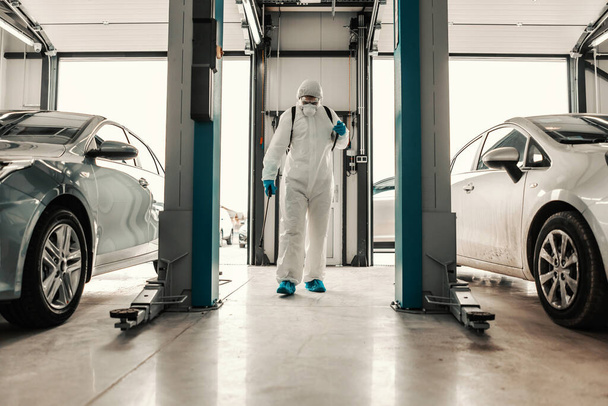 A male professional in a protective suit prevents the spread of the infection in the car garage. Coronavirus defense, stay healthy, warning for COVID 19, snitation, deep cleaning, pandemic situation - Foto, imagen