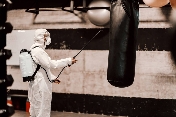 By disinfecting the space and air against the symptoms of coronavirus - fever, cough, headache. A man in a protective uniform cleans high-risk areas of infection such as a gym punching bag. COVID19 outbreak, keep social distance, coronavirus situatio - Foto, Bild