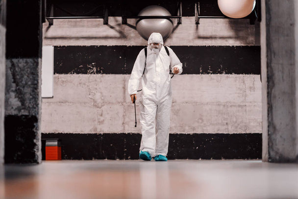 Warnung COVID 19. Ein Mann in weißer Schutzkleidung und ein Sprayer verhindern die Ausbreitung der Infektion in einem Flur des Fitness- und Pilates-Studios. Bleiben Sie verantwortlich, Coronavirus-Alarm - Foto, Bild