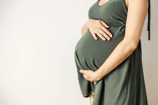 The pregnant woman's belly is in focus. The big belly of the future mother is in front of the white background. A woman in a casual gray-green dress stands and holds to her stomach. Motherhood - Foto, Bild