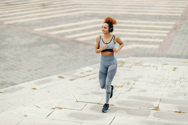 Gyors, fitt, göndör hajú, sportruhába öltözött sportnő fut az utcán fülhallgatóval a fején. Edzés közben a kedvenc rádióállomását hallgatja.. - Fotó, kép