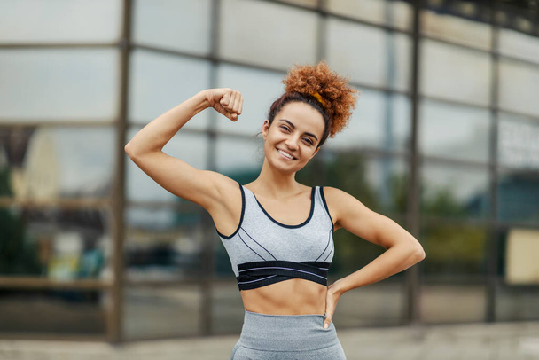 A young, strong, and happy sportswoman is showing her biceps while standing outdoors. Strength comes with effort and persistence, and the benefits are a happier and healthier life. - Fotografie, Obrázek