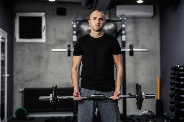 Proper posture before performing exercises. A man in sportswear pumps his arm muscles and lifts a barbell with the weights plate with both hands in a modern gym. A close-up shot of a male person - Photo, Image