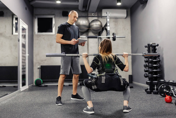 Une femme faisant de l'exercice dans une combinaison musculaire électrique stimule l'exercice dans une salle de gym. Une jeune femme en costume EMS tient une haltère sur ses épaules et fait des squats pendant que son entraîneur la surveille - Photo, image